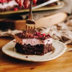 A piece of Chocolate Raspberry Brownie with pink marshmallow on top is being cut in to with a vintage fork, it is on a vintage plate, sitting on a wooden table.