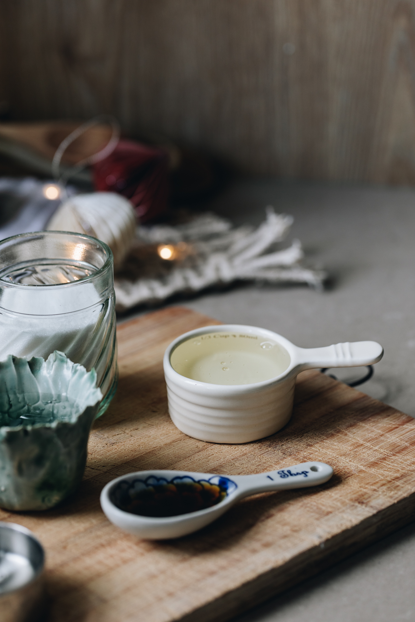 A wooden board sits on a stone bench. It has vintage spoons, cups and vases. They have glucose syrup, vanilla, water and sugar on it. A cream weaved mat is in the background and a soft glimmer of fairy lights. 