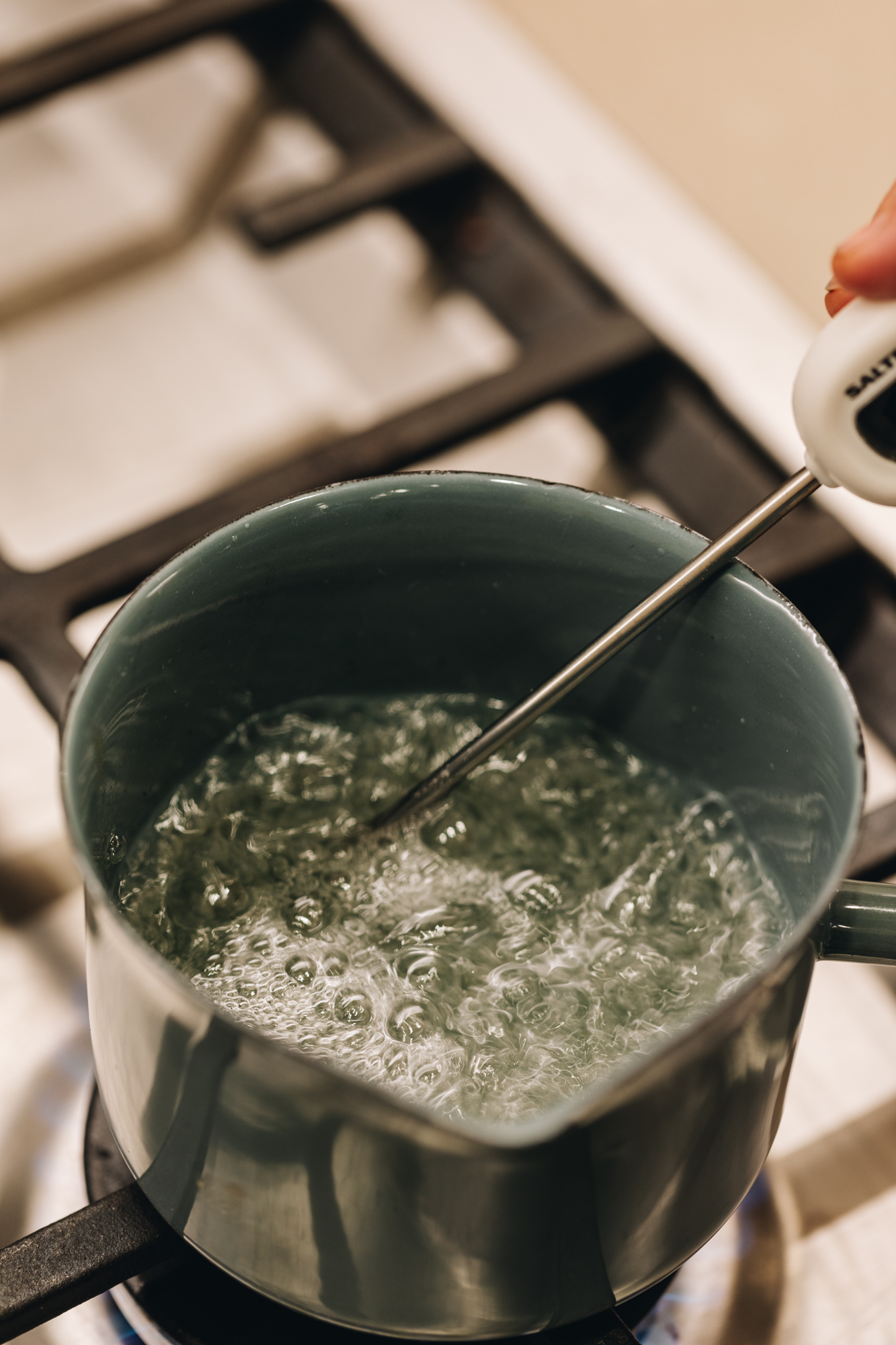 On a gas stove sits a small blue pot that has sugar syrup boiling in it, Naomi is holding a white handled sugar thermometer in the sugar syrup.