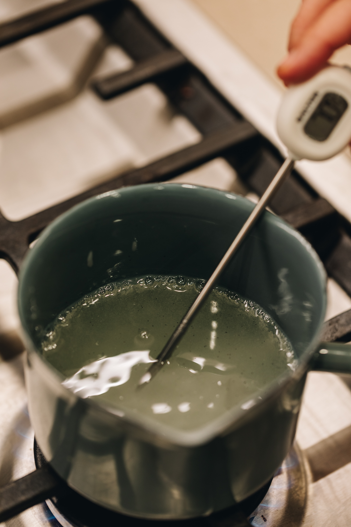 On a gas stove sits a small blue pot that has sugar syrup in it, Naomi is holding a white handled sugar thermometer in the sugar syrup.