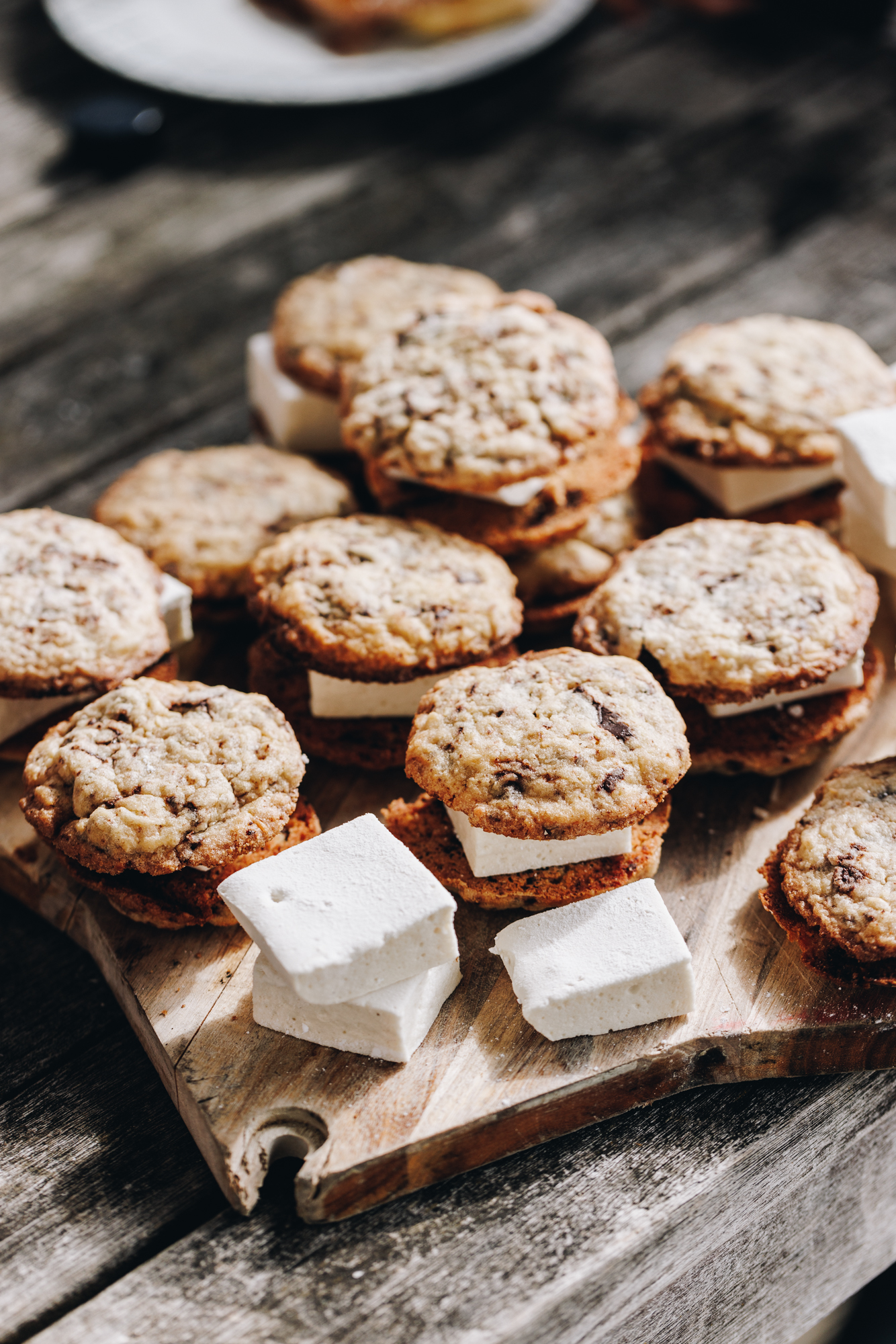 A wooden board sits on an old wooden deck. It is stacked with cookies and in-between each one is easy homemade vanilla marshmallows that has been cut in to squares. A plate is in the background.  