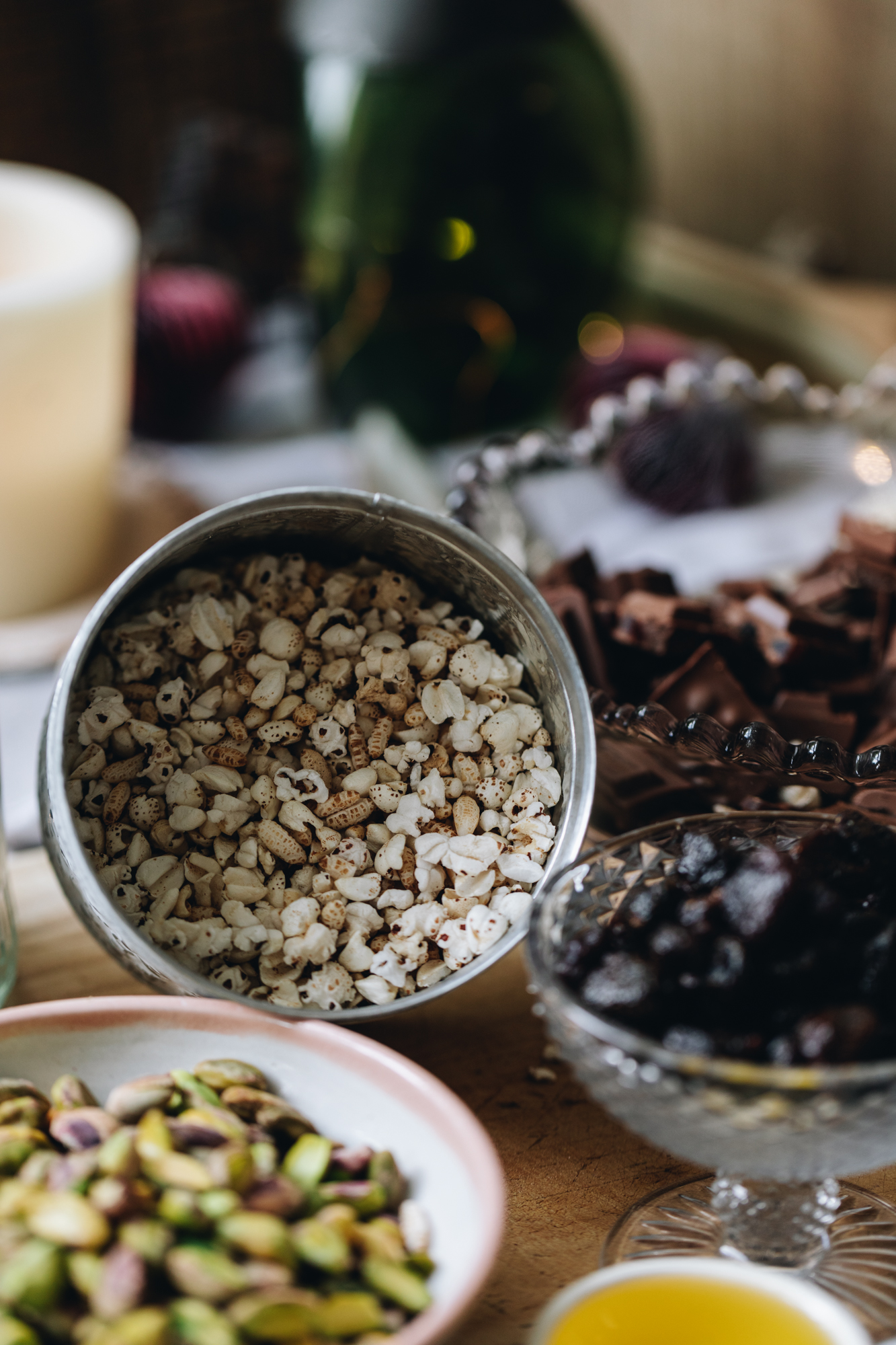 On a wooden board sits a white and silver jar with quinoa puffs in it, a white a pink bowl with pistachios, a glass vintage small bow with dried strawberries and a purple vintage bowl in the background has chocolate in it. In the far background is a candle burning with a green vase in the back. 