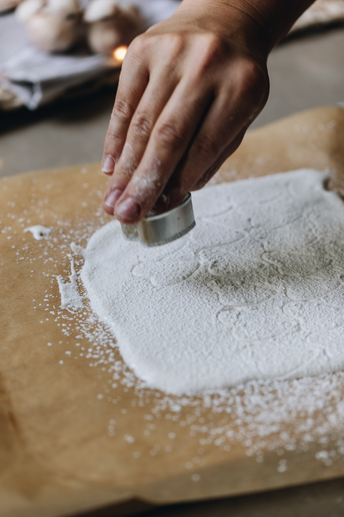 On brown baking paper is marshmallow that has been spread in a thin layer. A heart cookie cutter has cut out heart shapes all over it and a hand is cutting another one in to the marshmallow. It is sprinkled with cornflour. 