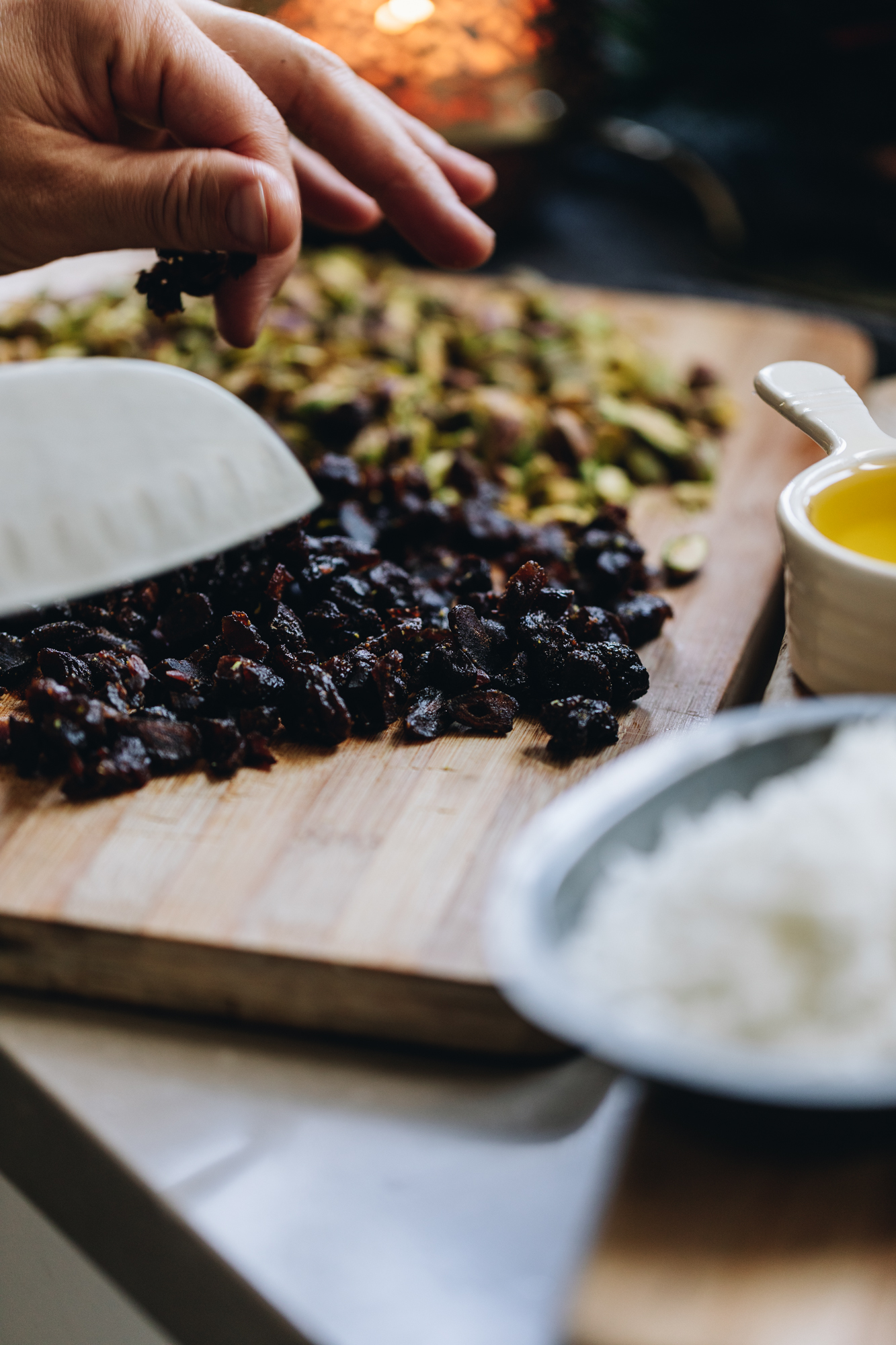 On a wooden board is chopped pistachios and cranberries. A small cup has oil in it. A candle burns in the background. 