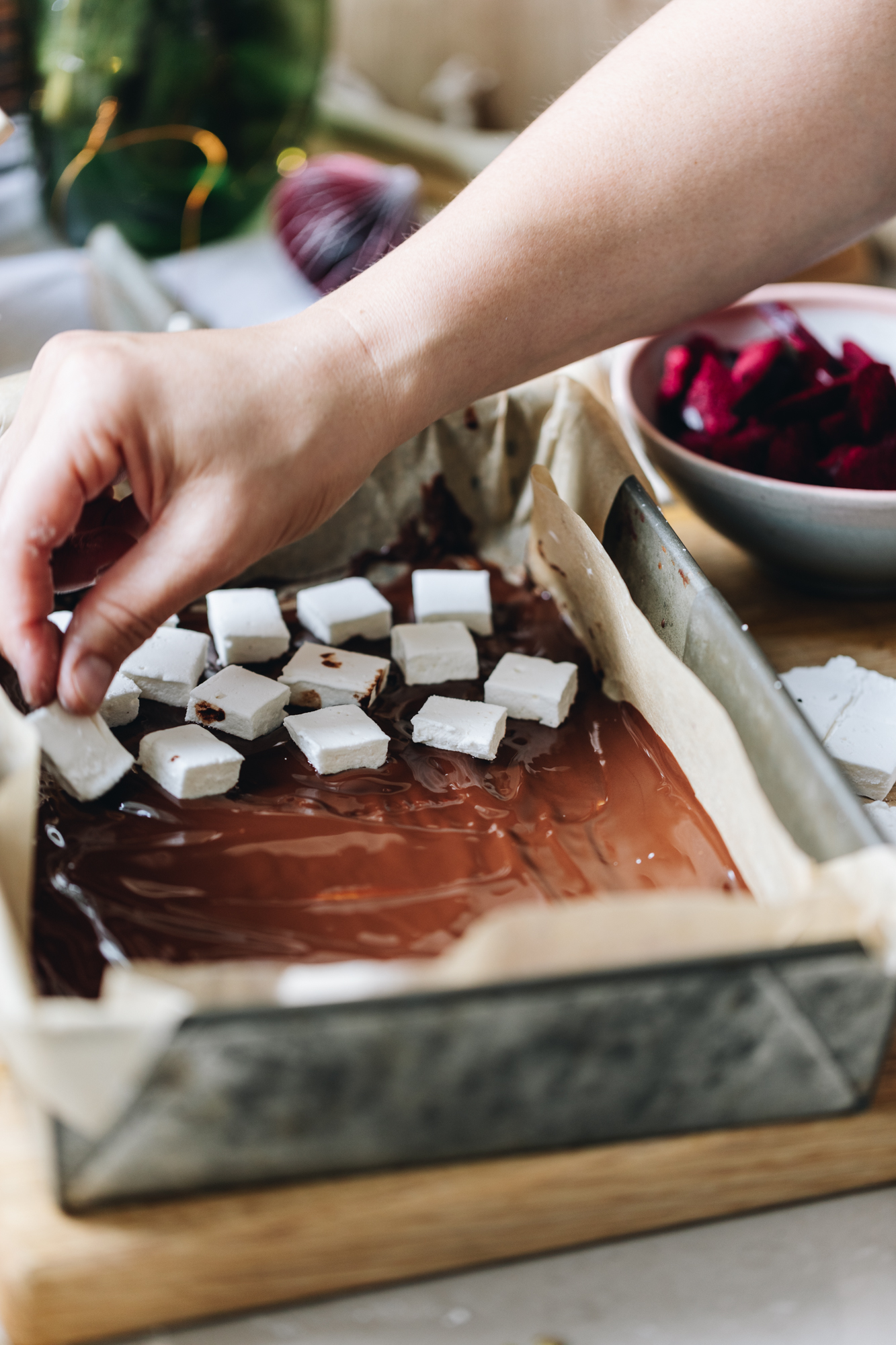 A baking tin lined with brown baking paper sits on a wooden board. It has dark chocolate being spread in to the base of the tin and squares of marshmallow cubes are being placed on top. Naomi is placing a piece of marshmallow on top od dark chocolate. In the background is a soft pink tablecloth and a bowl of freeze-dried plums. 