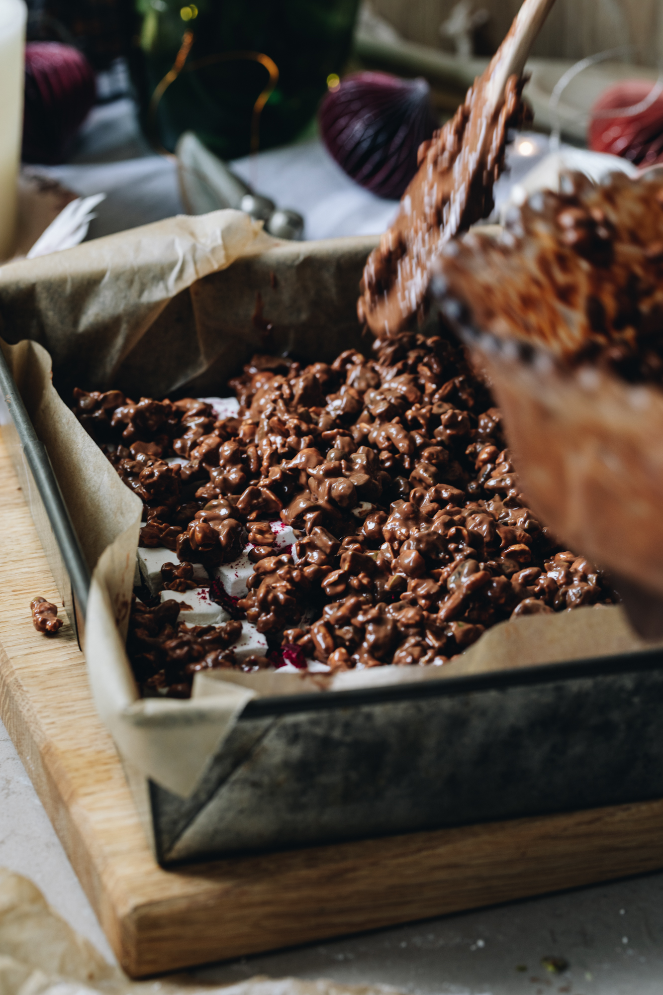 A baking tin lined with brown baking paper sits on a wooden board. It has dark chocolate being spread in to the base of the tin and squares of marshmallow cubes are placed on top. A milk chocolate mixture with nuts in it has been poured over the top. Th empty bowl is still in frame and a wooden spoon is hovering about the tin. This is the first layer of the homemade rocky road recipe.