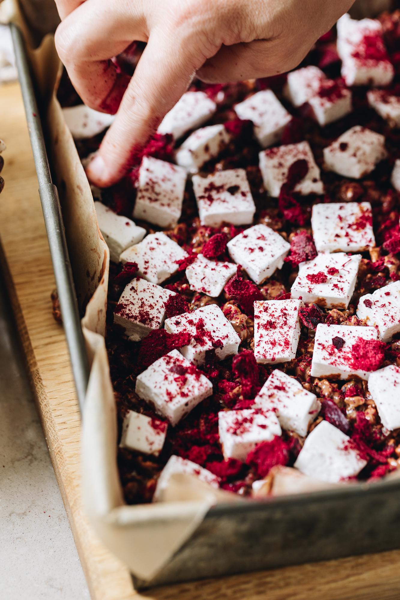 In a baking tray lined with baking paper is easy homemade vanilla marshmallow that has been cut in to squares and has freeze-dried plums sprinkled over top of it. Naomi's finger is pressing one of the pieces down. It is the second layer for the Homemade Rocky road recipe. 