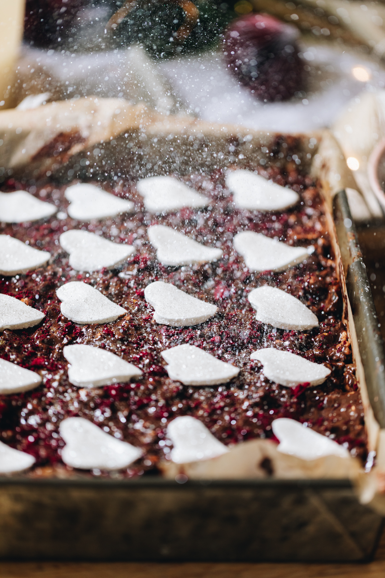 A baking tin is lined with borne baking paper and is filled with a homemade rocky road recipe. It has dark chocolate spread on top and crushed freeze-dried plums on top. Thin marshmallow heart sit on top of the rocky road and edible glitter is sprinkling down on top of it.