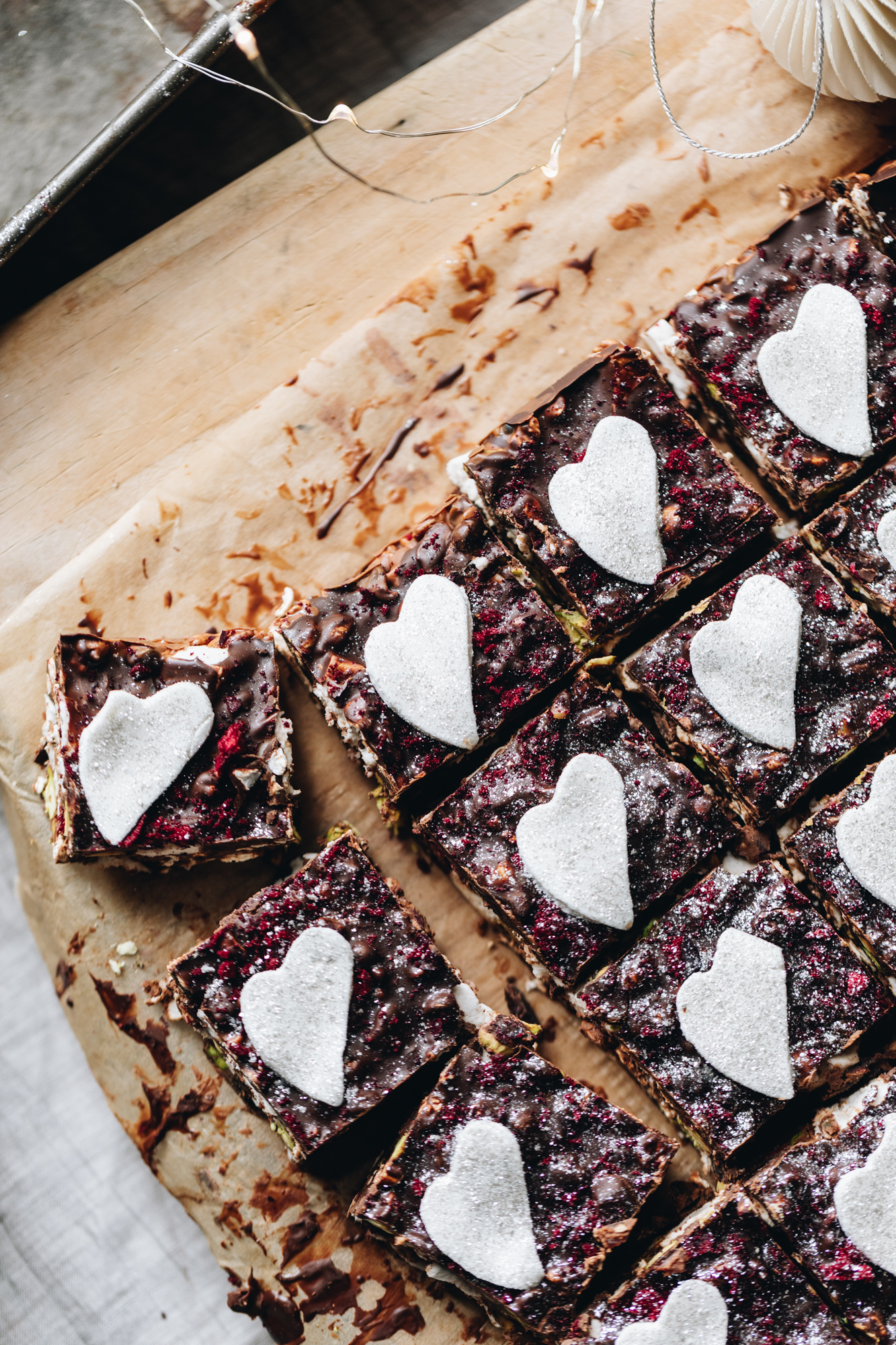 A flat lat shows a homemade rocky road recipe that has been cut in to pieces on top of brown baking paper, it is on a wooden board. It has dark chocolate spread on top and crushed freeze-dried plums on top. Thin marshmallow heart sit on top of the rocky road and edible glitter is sprinkled on top of it.