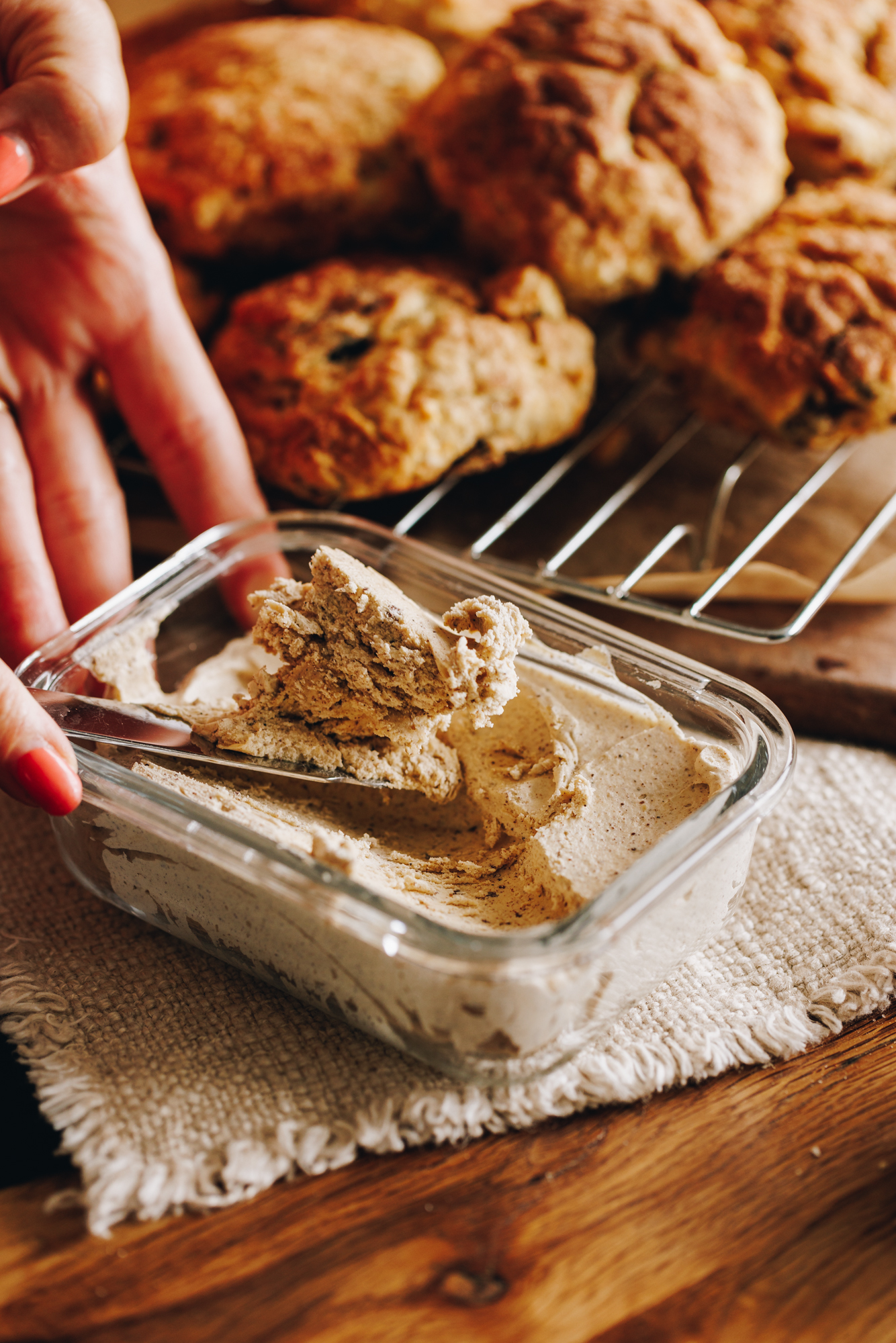 On a wooden table is a cream cloth that has a glass container filled with set whipped brown butter. A knife is digging out some. Behind it is a silver cooling rack with baked scones piles on top.