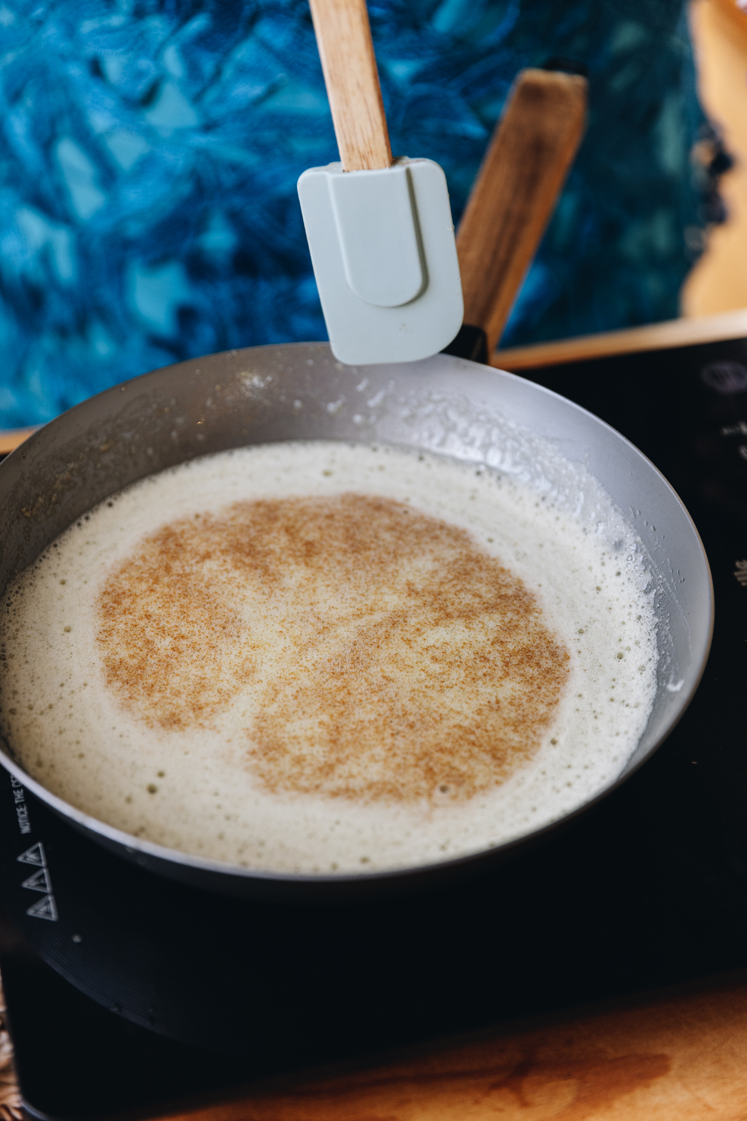 A silver pan with a brown handle sits on a black portable stove with brown butter that is foaming and brown bits have floated to the top. A blue spatula with a wooden handle is in shot and is about to stir it.