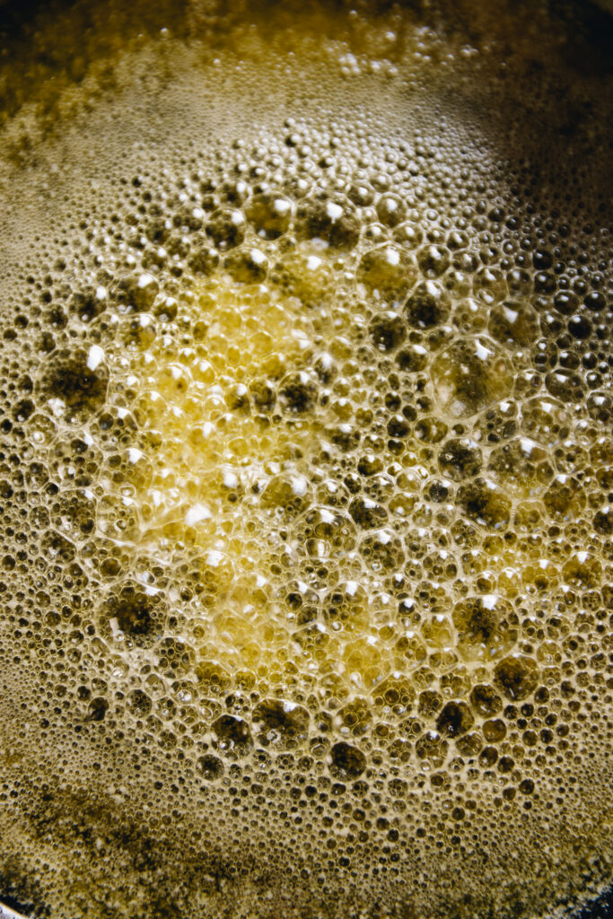 A close up shows butter that has clarified and the milk solids have separated and have gathered in the centre of the pan. The butter is foamy and bubbling.