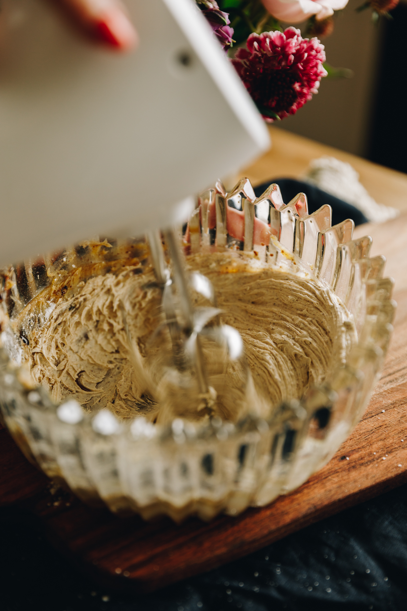 A glass vintage bowl sits on a wooden board. In the bowl is brown butter that is being whipped with a white hand mixer. Flowers are in the background.