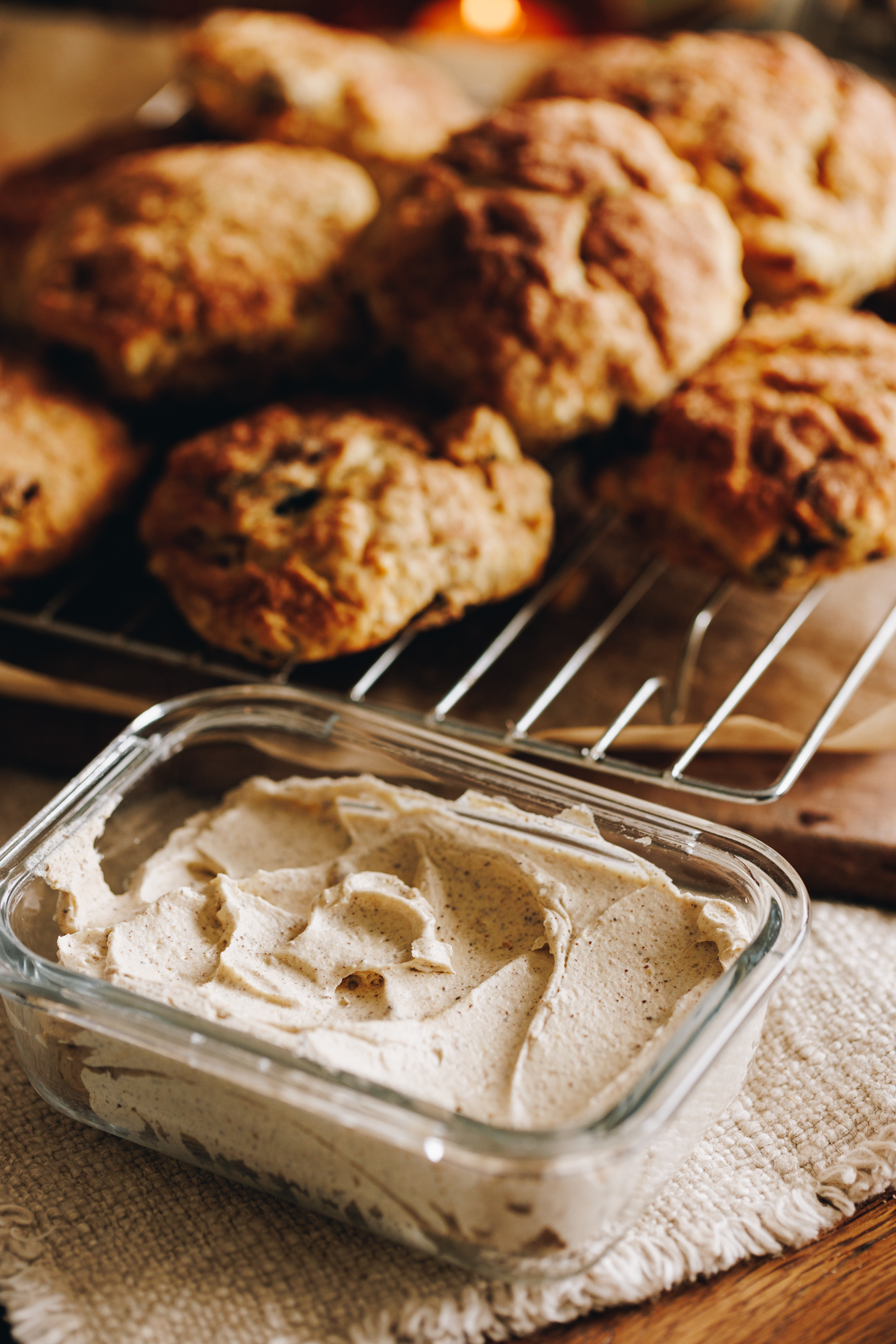 On a wooden table is a cream cloth that has a glass container filled with set whipped brown butter. Behind it is a silver cooling rack with baked scones piles on top.