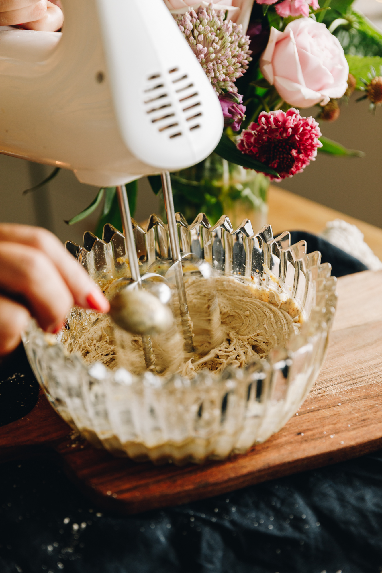 A glass vintage bowl sits on a wooden board. In the bowl is brown butter that is being whipped with a white hand mixer. Naomi is adding icing sugar in the bowl with a teaspoon as it whips. Flowers are in the background.