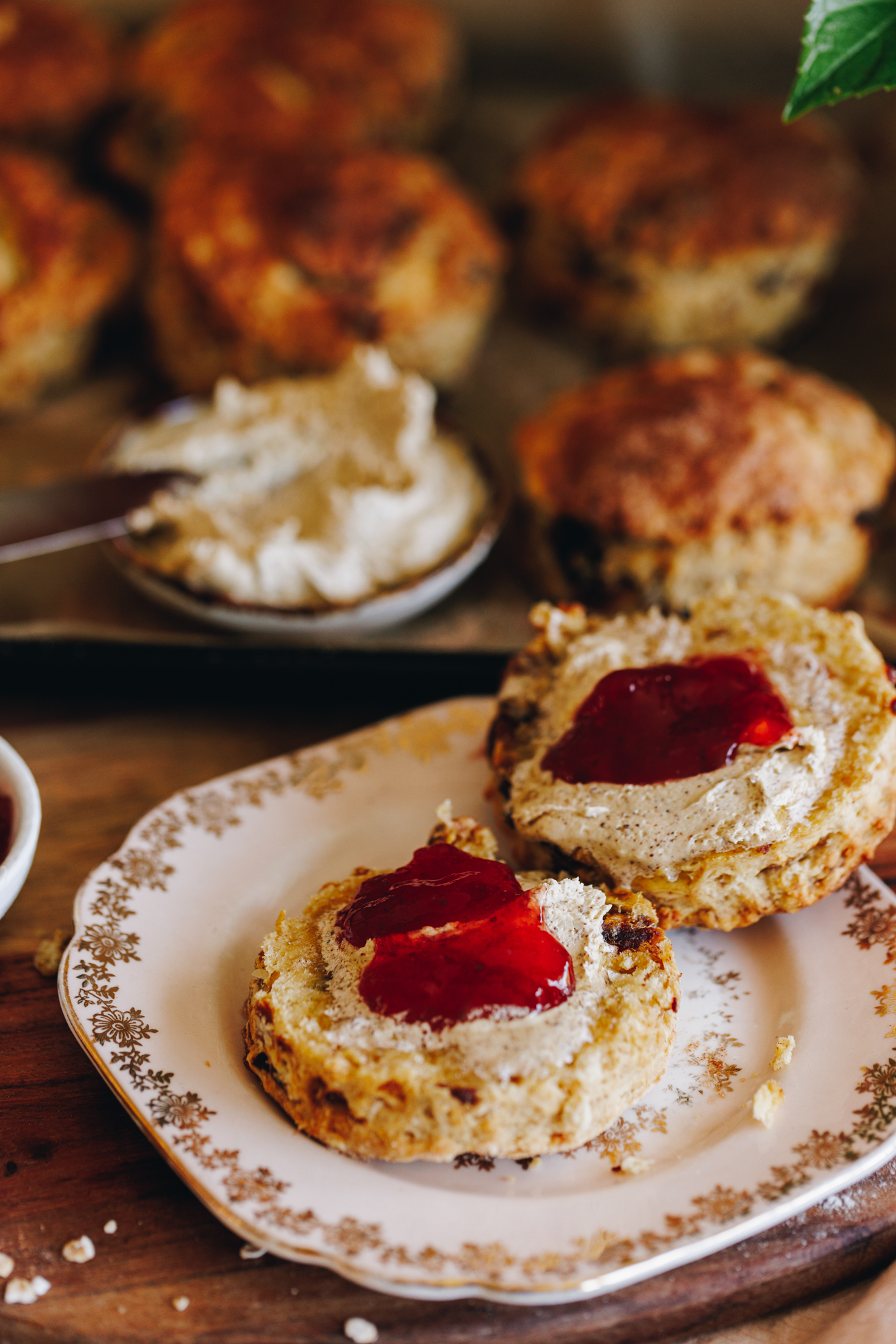 A pink vintage plate sits on a wooden table. On the plate is a date scone opened up with brown whipped butter on it and strawberry jam. Whipped butter and scones are in the background.