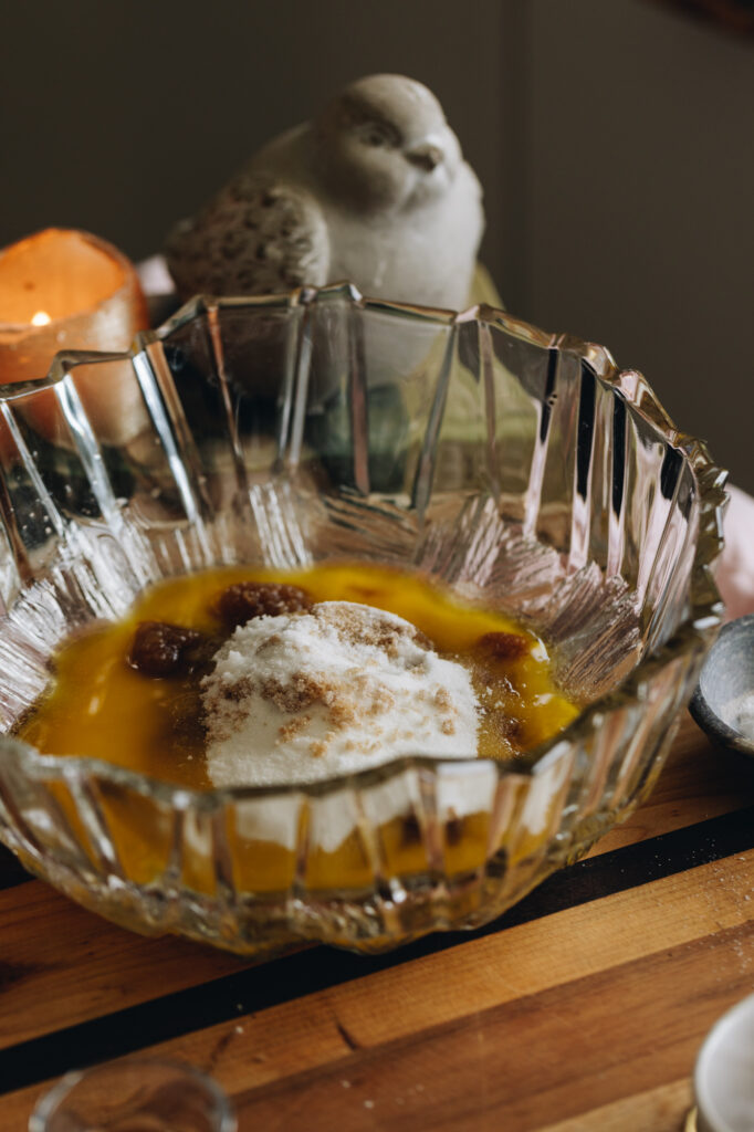 Melted butter and sugar are in a large glass bowl. It is sitting on a wooden board and a fat ceramic bird and a candle are in the background. 