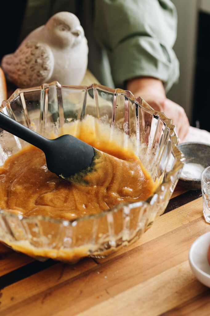 The eggs and butter mixture have been mixed for the Ultimate Peanut Butter Cookie. It is sitting on a wooden board and a fat ceramic bird and a candle are in the background. 