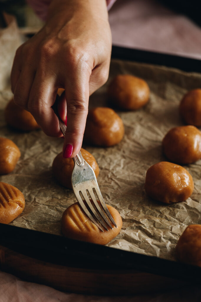 The cookie dough is unbaked and rolled in to balls. They are on a black tray with brown baking paper. One of the  cookies is being squashed with a vintage fork which makes grooves in the cookie. 