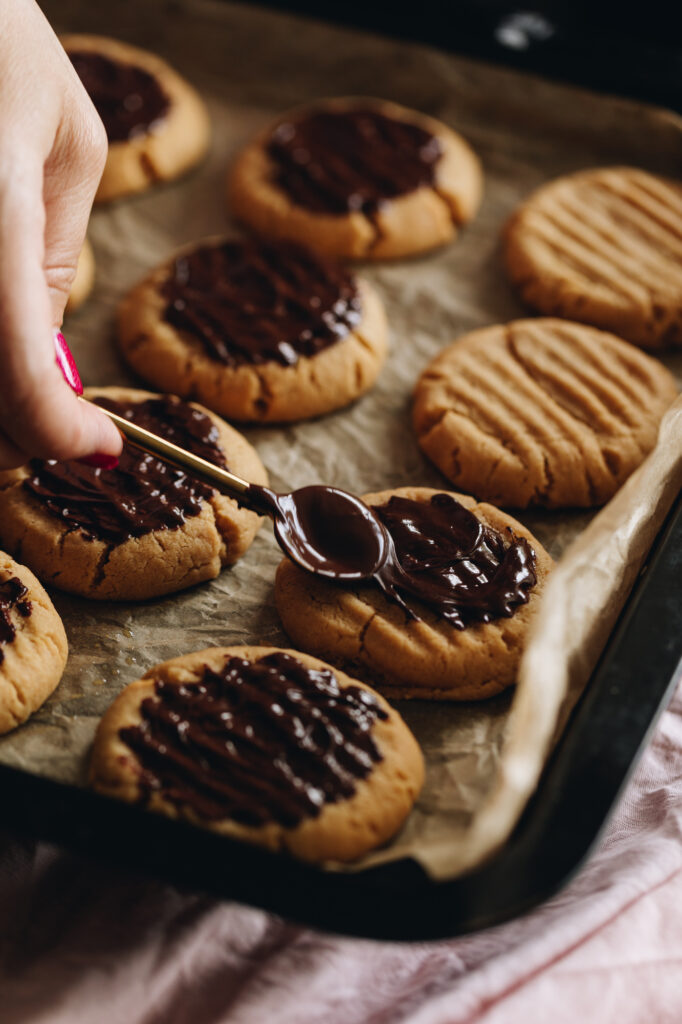 The baked cookies are on a black tray with brown baking paper. A teaspoon is smearing melted chocolate on to a cookie. Other cookies have the chocolate and others are plain. 