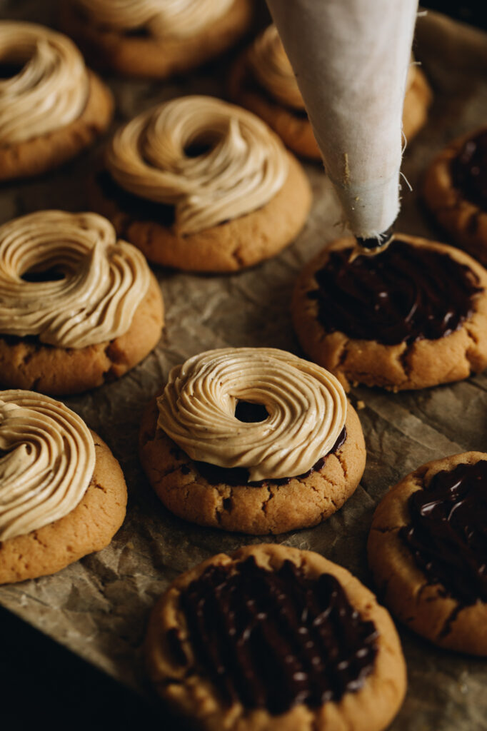 The baked Ultimate Peanut Butter Cookies are on a black tray with brown baking paper. Most of the cookies have been piped with peanut butter buttercream. A piping bag is suspended above one cookie, ready to pipe the next one. 