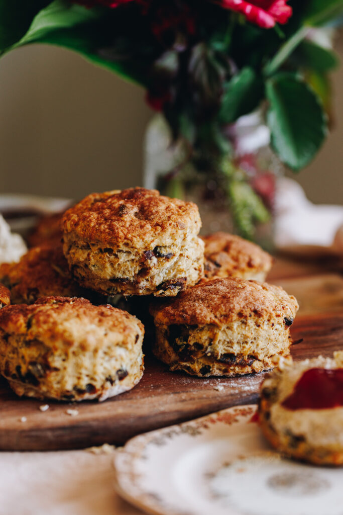 Baked Date and Orange Scones are in a pile on a wooden board. There is a glimpse of an open scone with butter and jam on it to the side of the photo. 