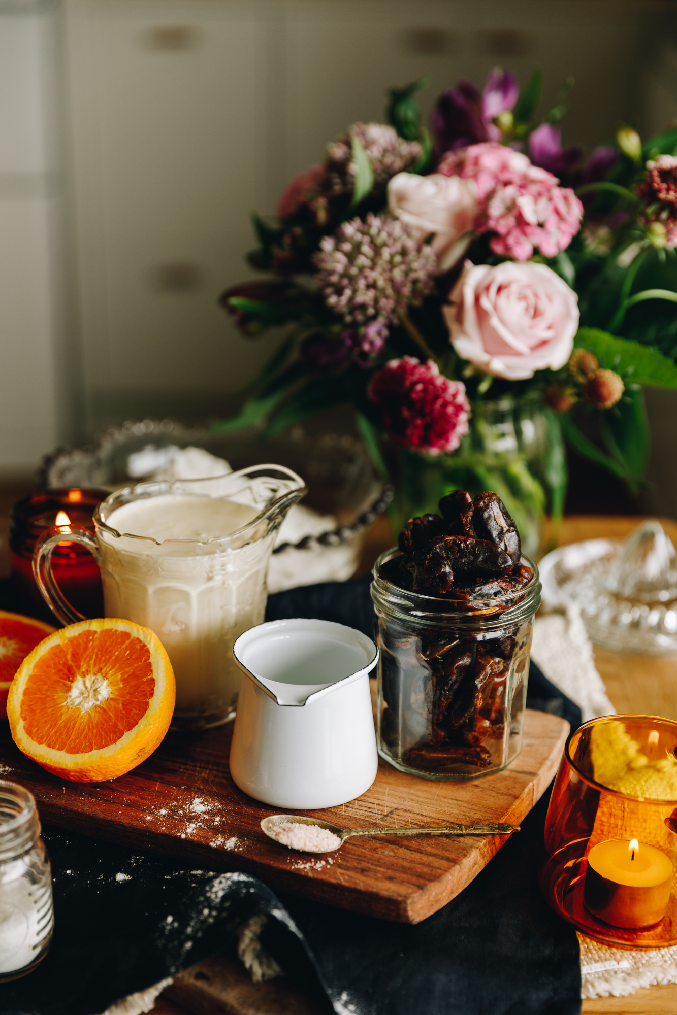 The ingredients of the Date and Orange Scones are in vintage glasses and jars. There is an orange sliced open and flowers in the background, with candles. 