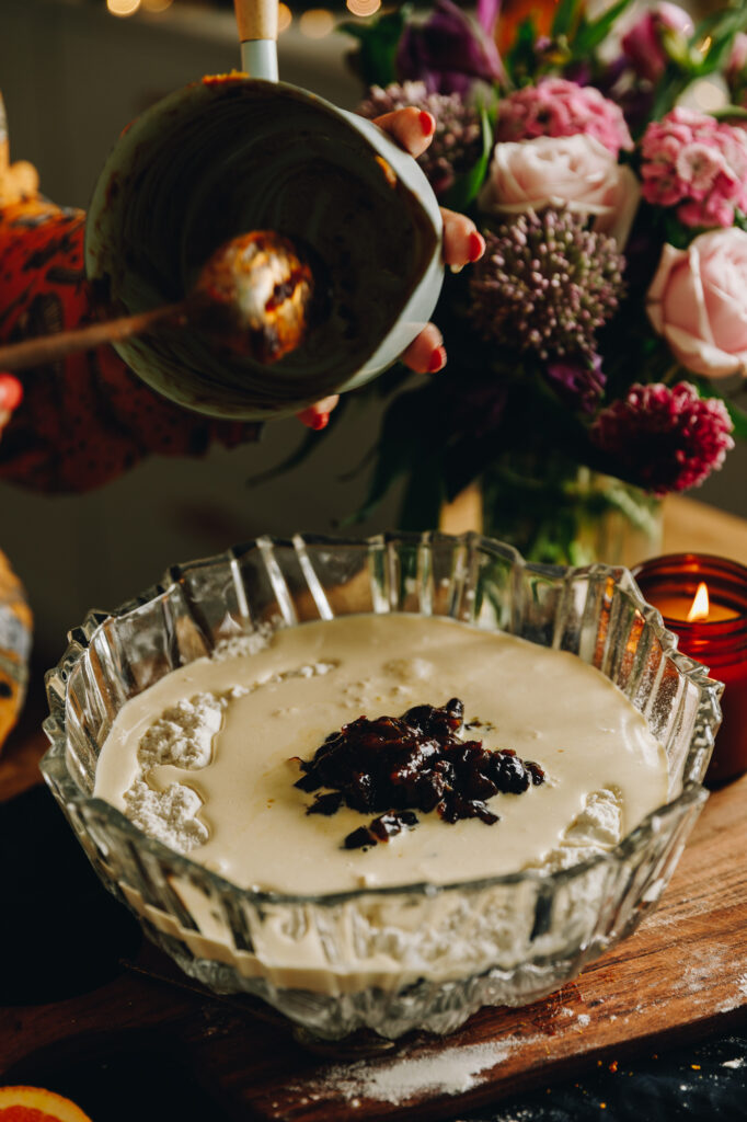 In a glass bowl is dry ingredients, runny cream and dates have been added from a small blue pot. It is sitting on a wooden board with a candle and flowers in the background. 