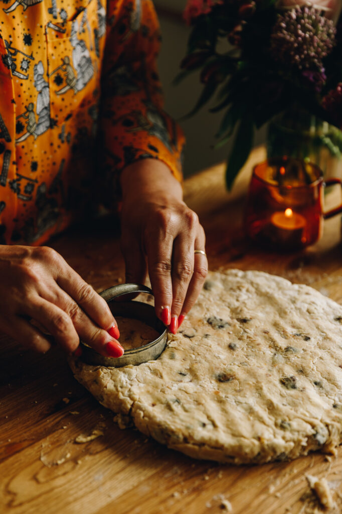 In a wooden table, Naomi Toilalo's hands are cutting the Date and Orange Scone dough with a cookie cutter. 