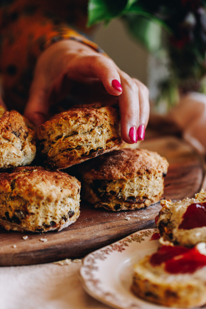 Baked Date and Orange Scones are in a pile on a wooden board. A hand is adding another scone in place. 