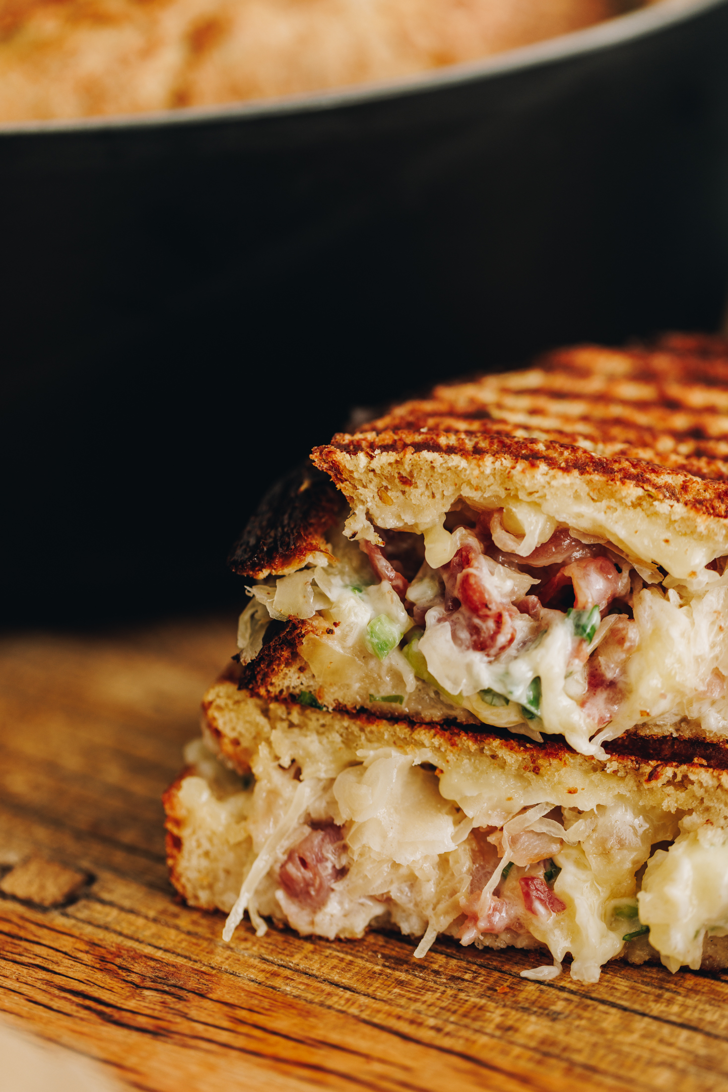 On a wooden board is freshly made maple bacon cheese toastie that is cut in half and stacked on top of each other. A black cat iron pot is in the background. The shot is super close revealing all of the bacon, cheese and sauerkraut.