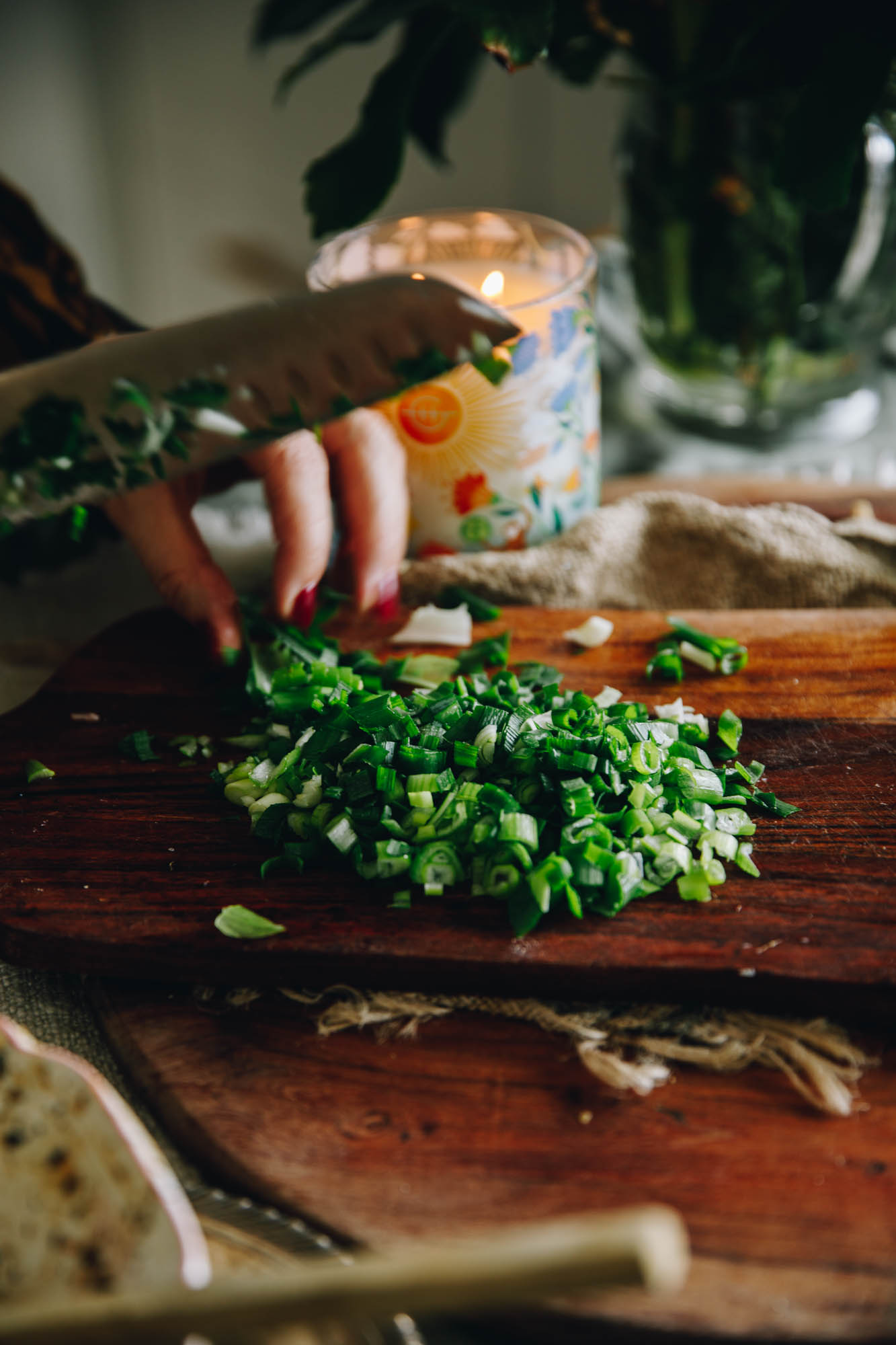 On a wooden board spring onions are being sliced. There is a blue candle in the background. It is sitting on another wooden board.