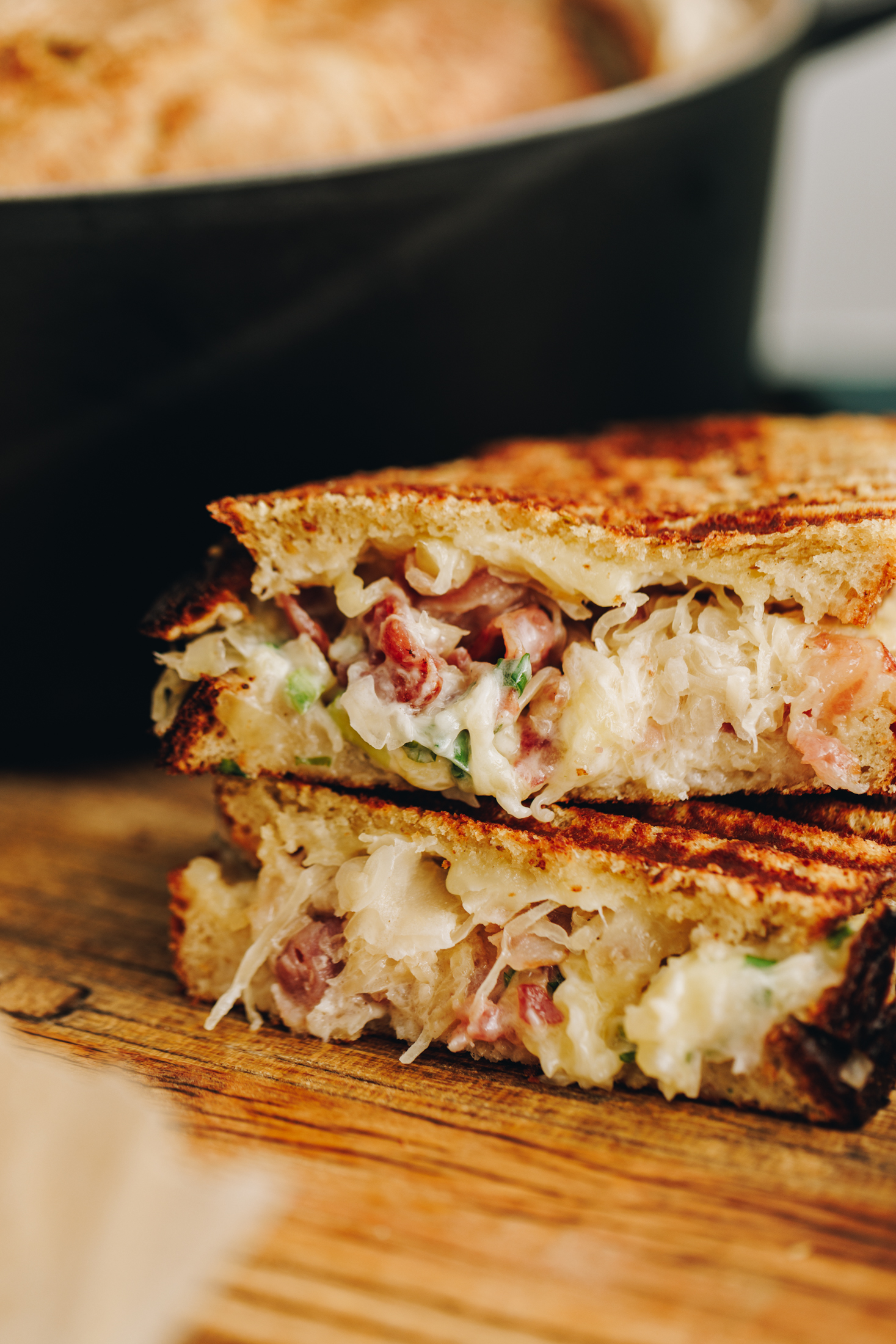 On a wooden board is freshly made maple bacon cheese toastie that is cut in half and stacked on top of each other. A black cat iron pot is in the background. The shot is super close revealing all of the bacon, cheese and sauerkraut.
