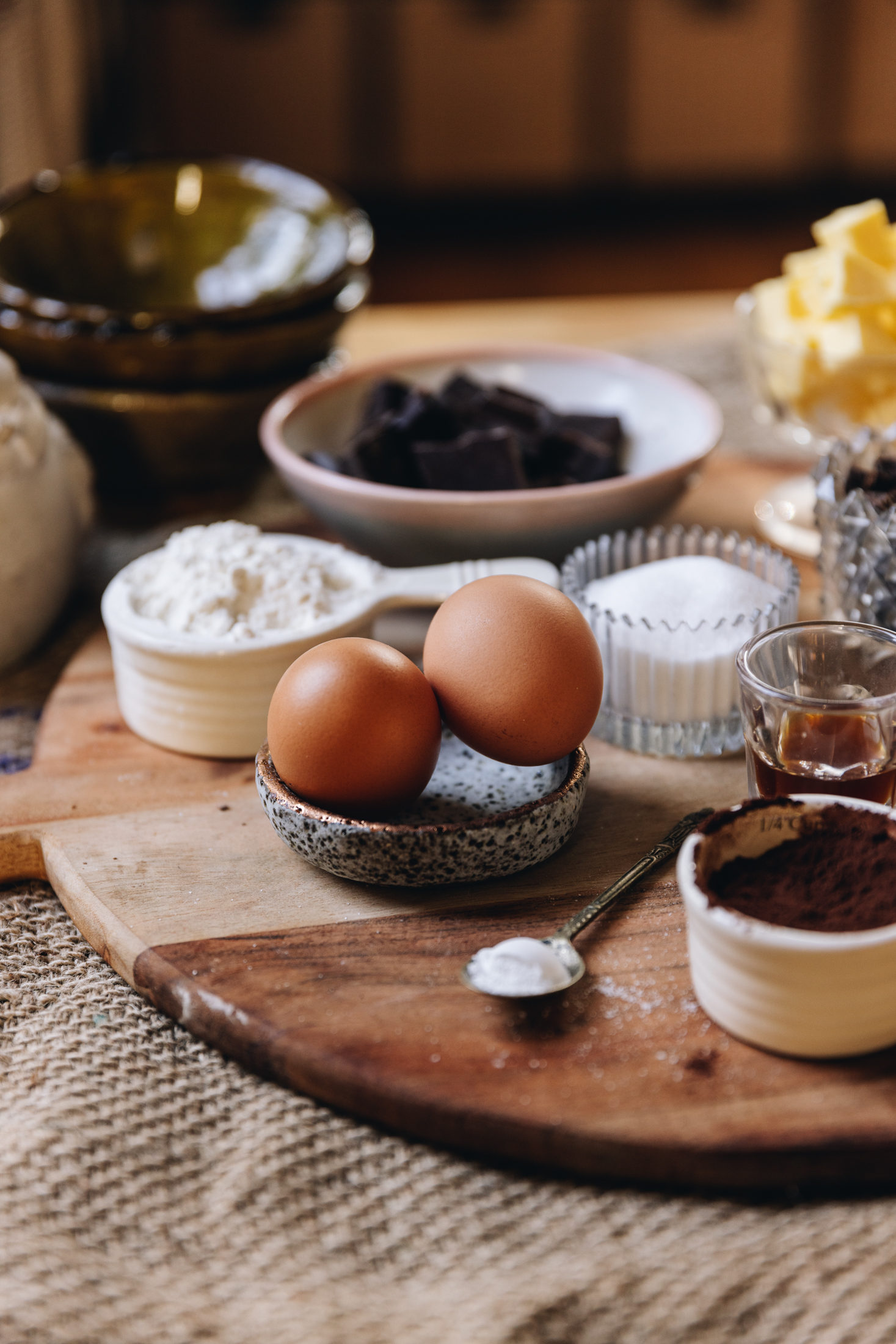 A wooden board is filled with biscuit ingredients and is sitting on natural fabric. There is eggs, cocoa, chocolate, sugar and butter in shot. A stack of ceramic bowls and a ceramic bird are in the background.