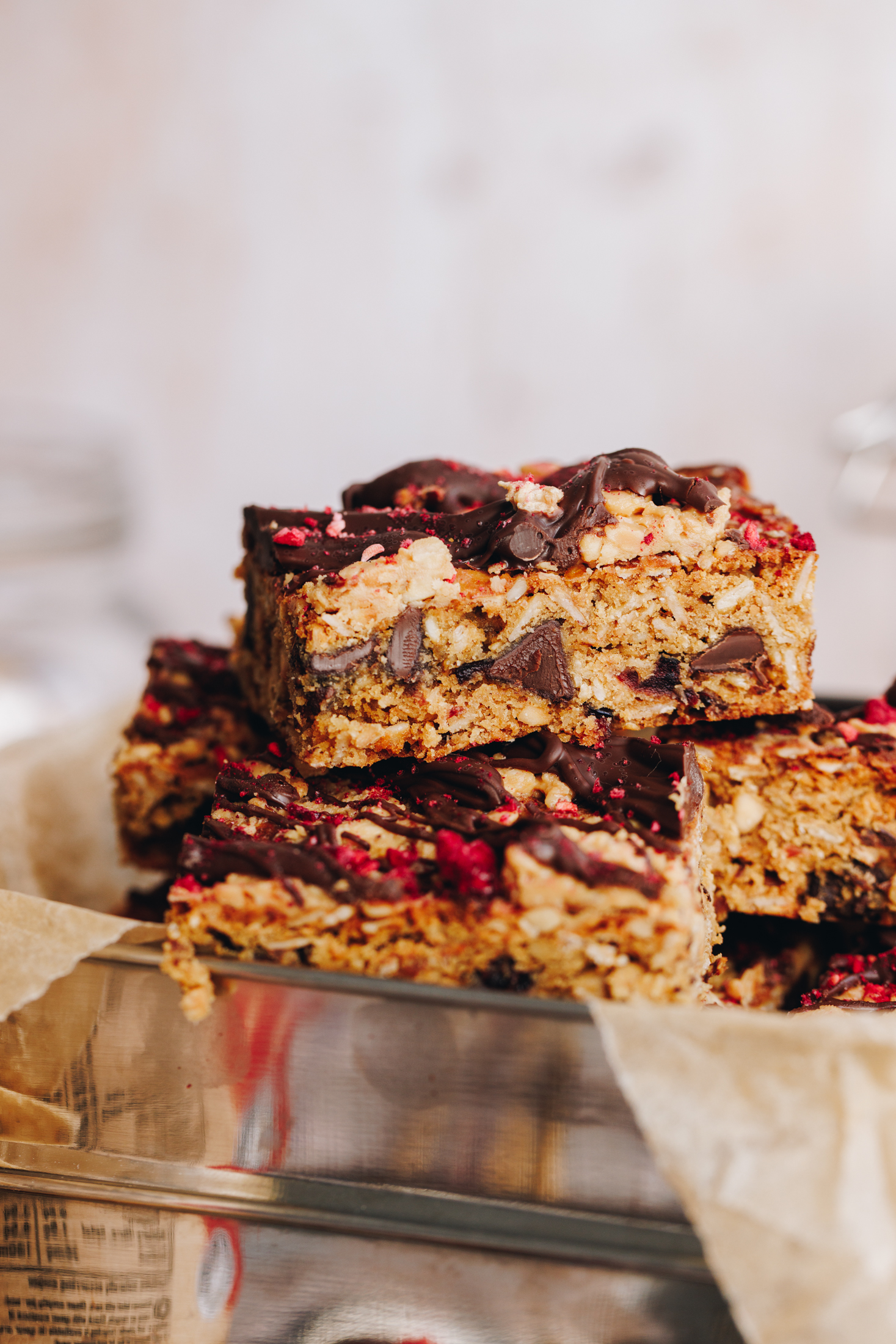 Peanut butter muesli bars are sliced up and are on stacked on top of each other. They are in a silver lunchbox that is lined with brown baking paper. The bars are golden and have chunks of chocolate  in the them. They have chocolate and freeze-dried raspberries set on top. 