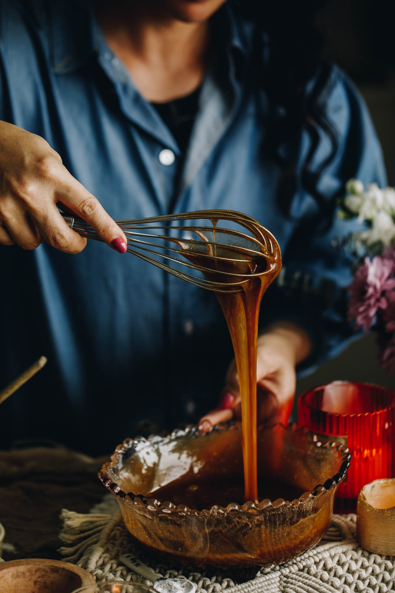 A purple vintage mixing bowl is sitting on a weaved creamed mat, on a wooden table. In the bowl is whipped egg and butter mixture. Naomi is holding a whisk above the bowl and the mixture is dripping off it.