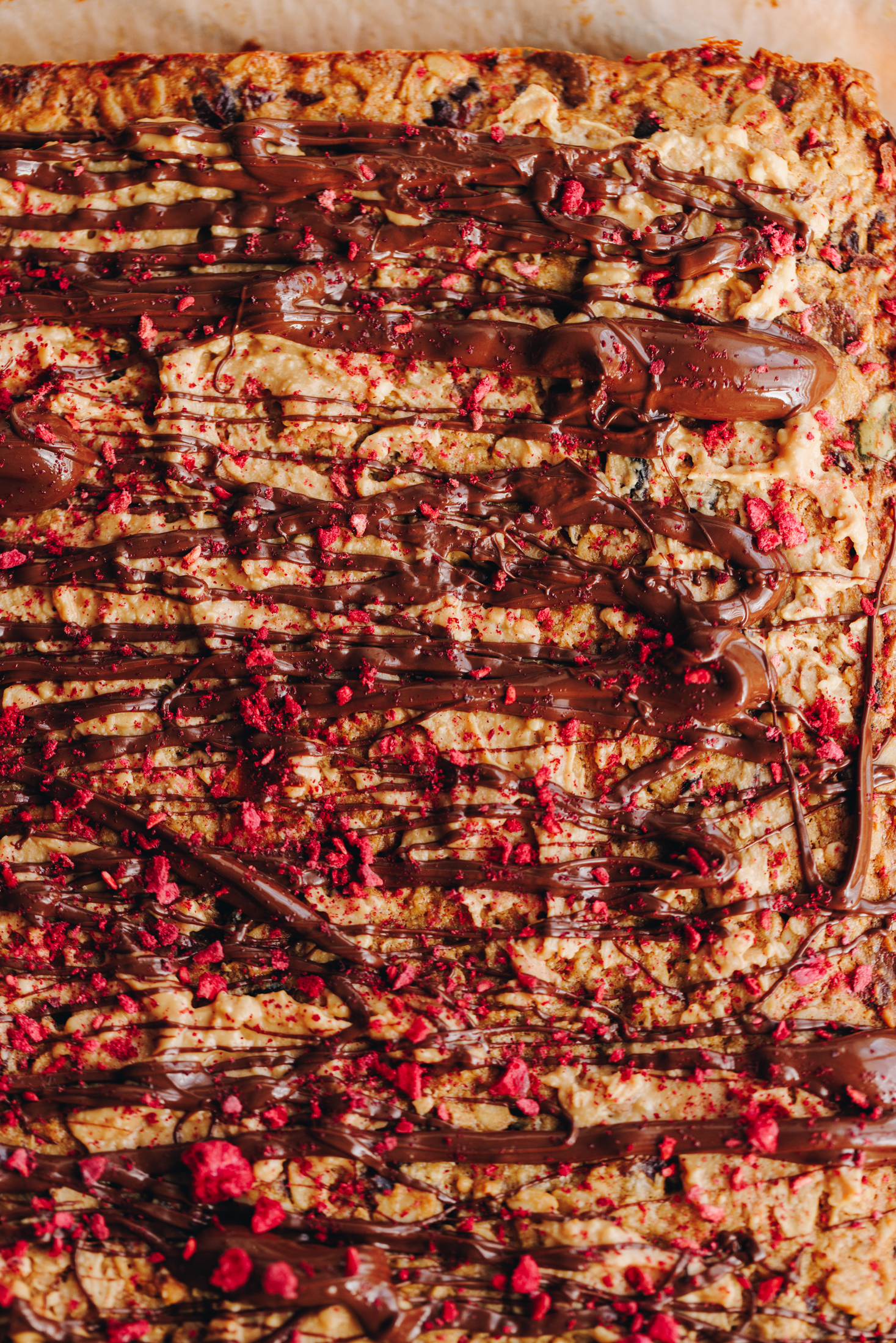 A flat lay shot of the baked peanut butter muesli bars reveal a golden baked slice with melted dark chocolate drizzled on it with a sprinkle of freeze-dried raspberries. 