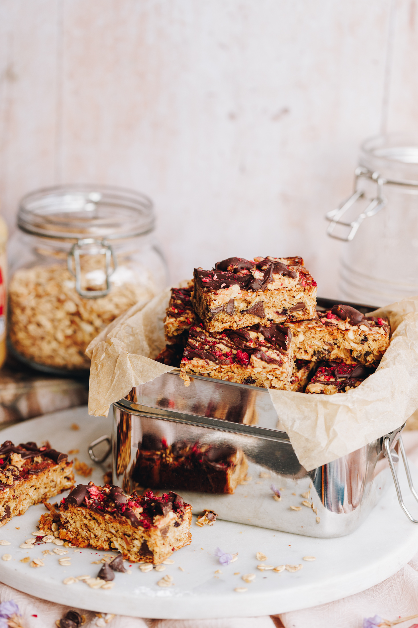 Peanut butter muesli bars are sliced up and are on stacked on top of each other. They are in a silver lunchbox that is lined with brown baking paper and is sitting on a white, round stone. The bars are golden and have chunks of chocolate in the them. They have chocolate and freeze-dried raspberries set on top. There are jars with oats and ingredients in the background and a couple of slices are next to the lunch box on the white stone. 