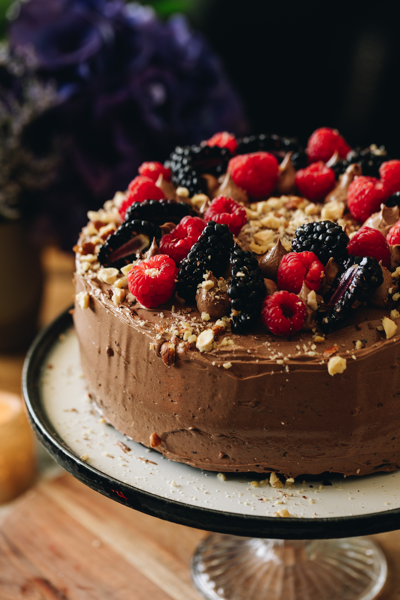 A black and white plate sits on a glass cake stand, it sits on a wooden board. On the plate is a chocolate buttercream covered gluten-free chocolate hazelnut cake. The topping has crushed hazelnuts, fresh blackberries and fresh raspberries arranged on top.