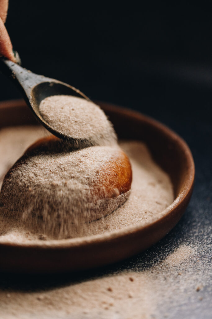 A freshly fried Chocolate Stuffed Fry Bread is in a bowl of cinnamon sugar and a wooden spoon is adding cinnamon sugar on top of the fry bread. 