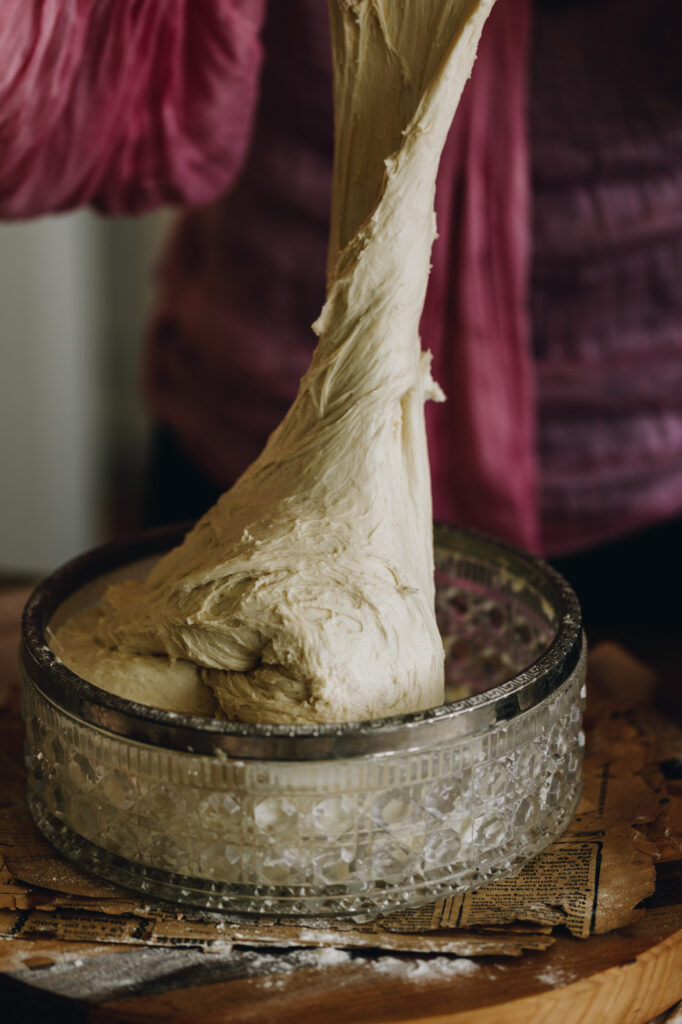 A dough is being stretch high in to the air out of a glass bowl with a silver trim. The dough looks stretchy.