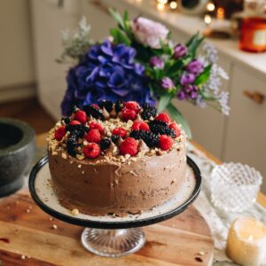A whole decorated Gluten-free Chocolate Hazelnut Cake is on a plate, on a cake stand that is on a wooden board. There are raspberries, blackberries and hazelnut crumbs on top. Flowers are in the background.
