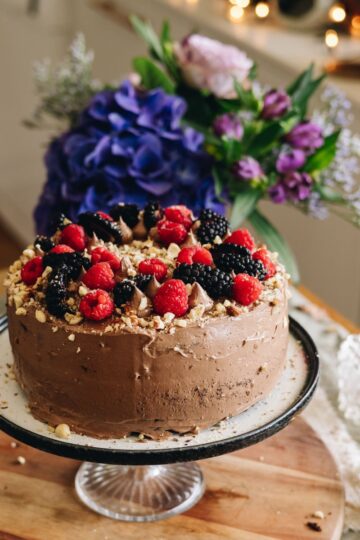 A whole decorated Gluten-free Chocolate Hazelnut Cake is on a plate, on a cake stand that is on a wooden board. There are raspberries, blackberries and hazelnut crumbs on top. Flowers are in the background.