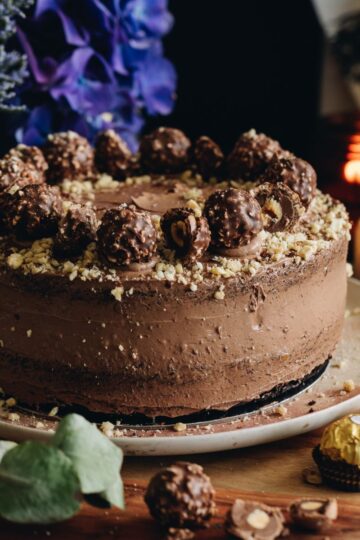 A whole decorated chocolate Hazelnut cake is on a pink plate, sitting on a wooden board. It is decorated with chocolate buttercream and forerro rocher chocolates.