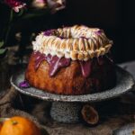 A Lemon Syrup Bundt Cake is a on a ceramic cake stand. It has drizzle boysenberry ganache drizzling down off the cake and is topped with swirly torched meringue.