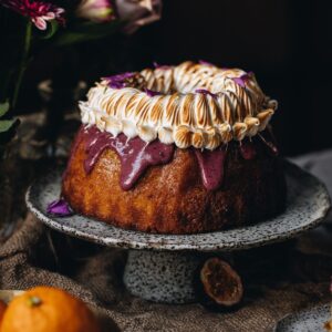 A Lemon Syrup Bundt Cake is a on a ceramic cake stand. It has drizzle boysenberry ganache drizzling down off the cake and is topped with swirly torched meringue.