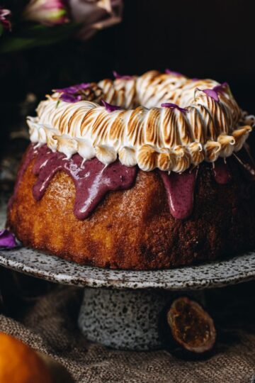 A Lemon Syrup Bundt Cake is a on a ceramic cake stand. It has drizzle boysenberry ganache drizzling down off the cake and is topped with swirly torched meringue.
