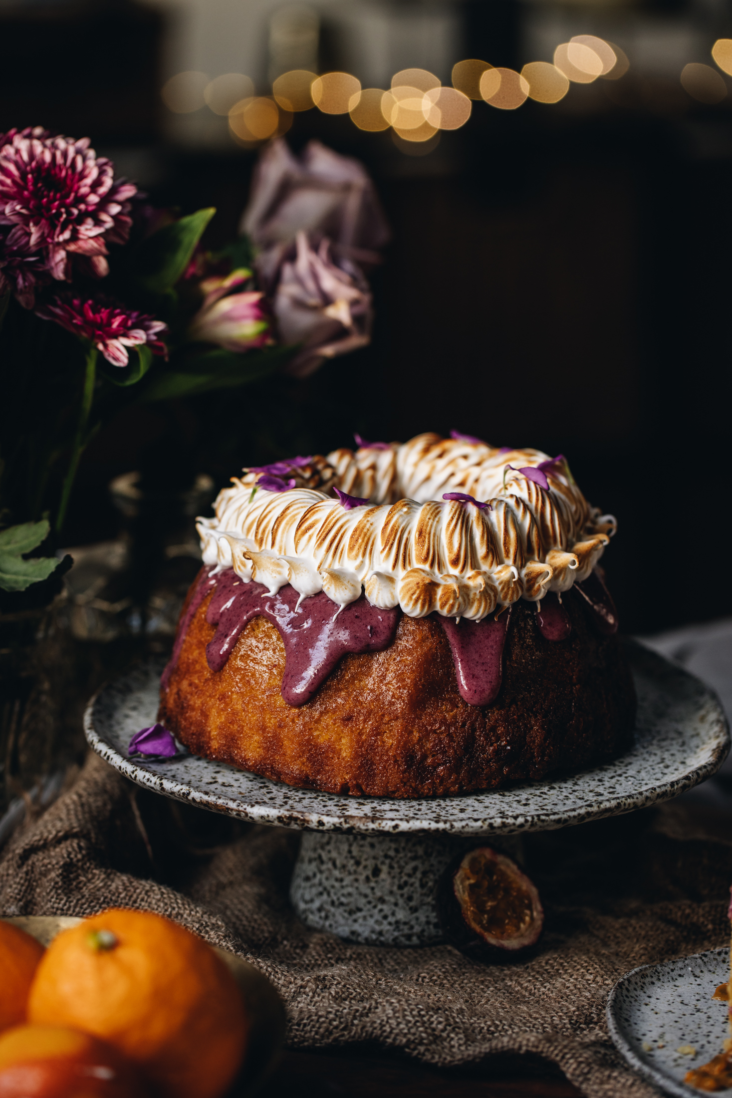 Sitting on natural fabric is a ceramic cake stand with a lemon syrup bundt cake on top of it. It is decorated with a boysenberry ganache dripping down the sides and is topped with piped meringue that has been torched. There are flowers behind it and glowing fairy lights.
