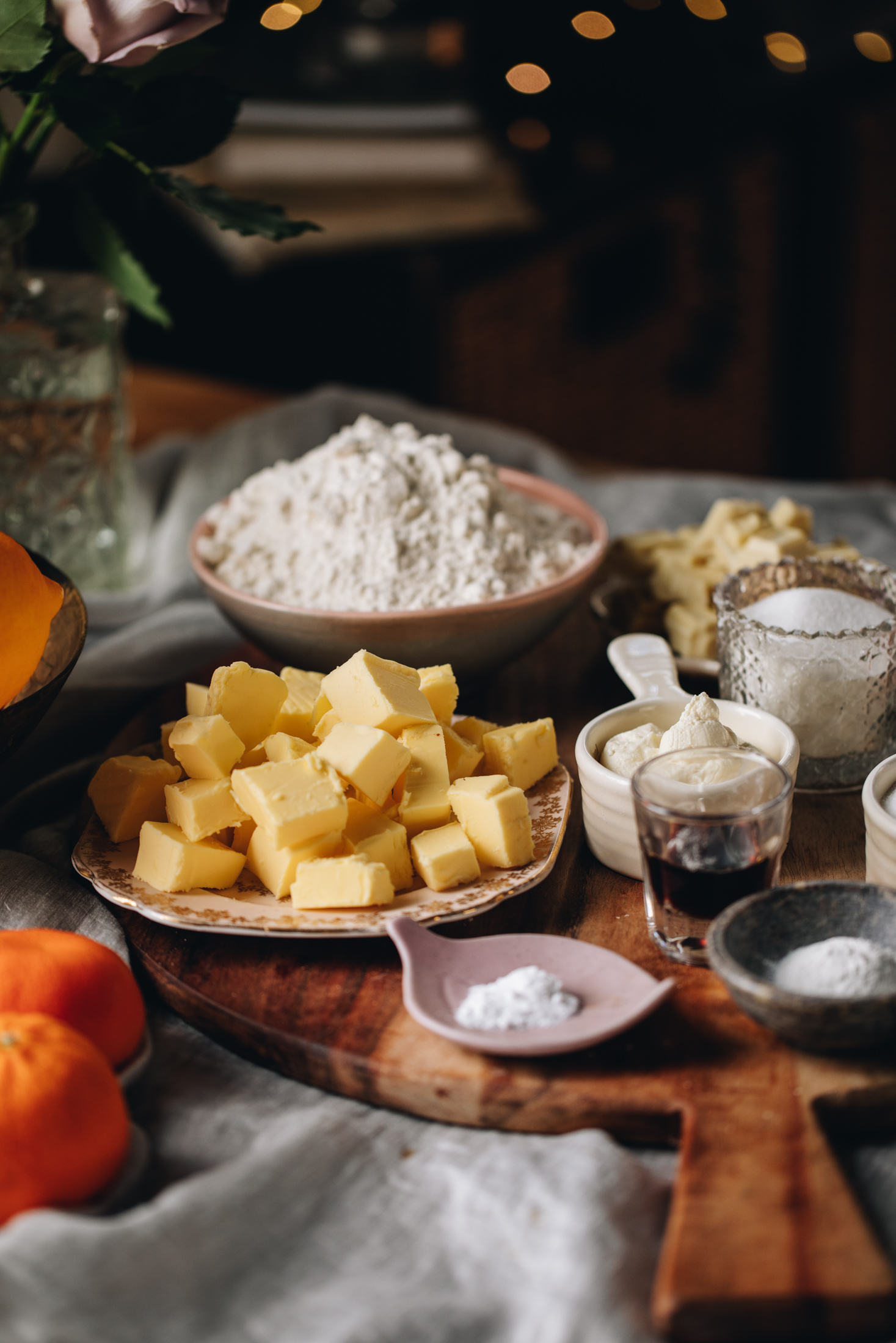 On a table is a soft grey tablecloth with a wooden board on it. On the board is vintage plates and cups with cake ingredients on it. There is butter, flour, baking powder, mandarins and vanilla in view. Soft fairy lights are seen in the background. 