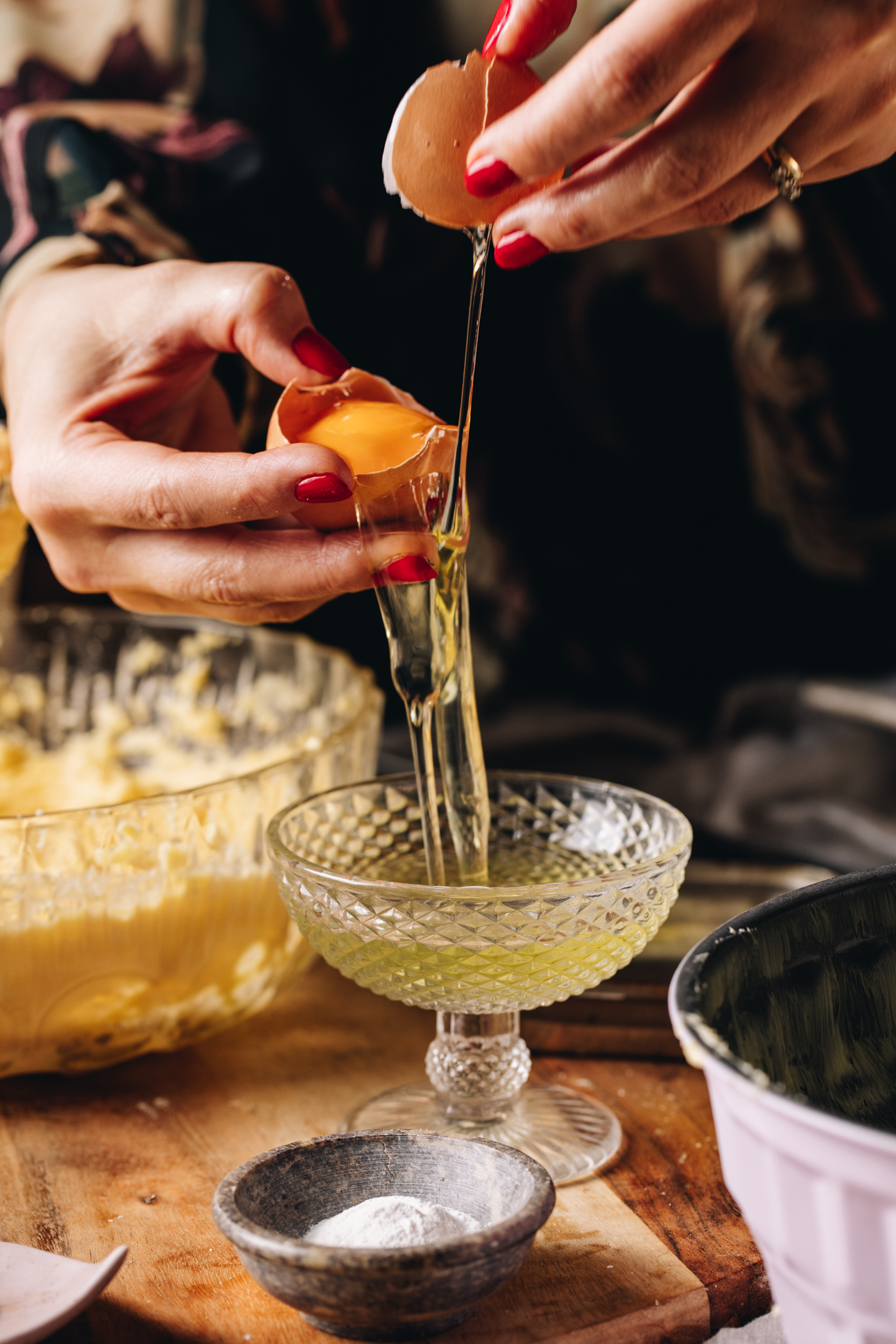 A small glass bowl with a stand is on a wooden bench and Naomi is separating and egg in to it. The egg yolk is one part of the egg and the whites are going in to the bowl. A large bowl of whipped butter is in the background. 